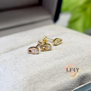 May include: Three gold earrings with a teardrop design, each featuring a small, clear gemstone. The earrings are displayed on a grey surface, with a blurred green background. The earrings are in different shades of gold.