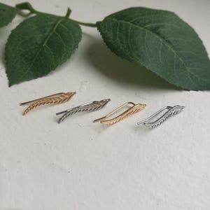May include: Four ear climbers in gold and silver tones, each featuring a delicate leaf design. The earrings are arranged on a white surface with green leaves in the background, creating a natural aesthetic.