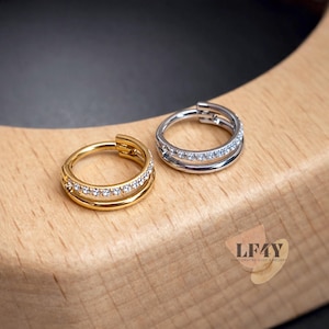 May include: Two hinged body jewellery rings, one gold-toned and one silver-toned, each with a double row of small, clear gemstones. The gold ring is on the left, and the silver ring is on the right. The jewellery is displayed on a light-coloured wooden surface.