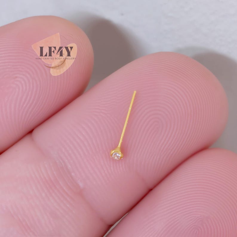 K&ouml;nnte beinhalten: Ein goldfarbener Nasenstecker mit einem kleinen, klaren Edelstein am Ende des Stifts. Der Stecker wird zwischen zwei Fingern gehalten. Das Bild zeigt auch einen Aufkleber mit dem Text "LF4Y HAND CRAFTED BODY JEWELLERY".