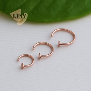 Puede incluir: Se muestran tres aros de nariz de oro rosa sobre un fondo suave. Cada aro tiene un diseño curvo con un pequeño disco plano en un extremo. Los aros son de diferentes tamaños, mostrando un estilo minimalista y elegante. El logotipo "LF4Y Crafted Body Jewellery" está en la esquina superior izquierda.