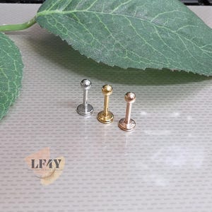 Może przedstawiać: Trzy kolczyki do ciała w stylu labret w kolorze srebrnym, złotym i różowego złota. Każdy kolczyk ma kuliste zakończenie i płaskie, dyskowe oparcie. Biżuteria jest prezentowana na jasnej powierzchni z zielonym liściem w tle.