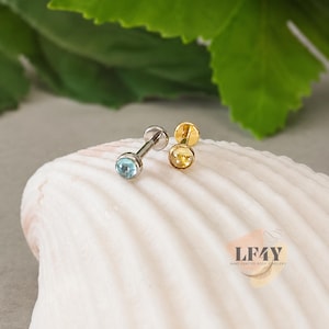 Op de afbeelding: Twee kleine oorstekers. Eén is zilverkleurig met een lichtblauwe edelsteen en de andere is goudkleurig met een gele edelsteen. De oorbellen worden gepresenteerd op een witte zeeschelp, met groene bladeren op de achtergrond.