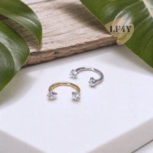 May include: Two circular horseshoe-shaped body jewellery pieces, one in gold tone and one in silver tone, each featuring a clear round-cut gemstone at both ends. The text LF4Y is visible in the upper right corner of the image.