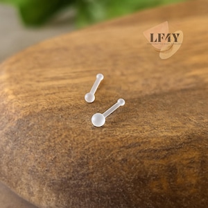 Könnte beinhalten: Zwei kleine, klare Nasenknochen mit abgerundetem Ende und geradem Stab. Der Schmuck wird auf einer Holzoberfläche präsentiert. Das Logo "LF4Y HAND CRAFTED BODY JEWELLERY" befindet sich in der oberen rechten Ecke.