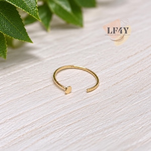 Puede incluir: Un aro nasal abierto de color dorado con un pequeño disco circular plano en un extremo. La joya se muestra junto a un logotipo que dice LF4Y HAND CRAFTED BODY JEWELLERY. El aro tiene una estructura de alambre circular y fina.