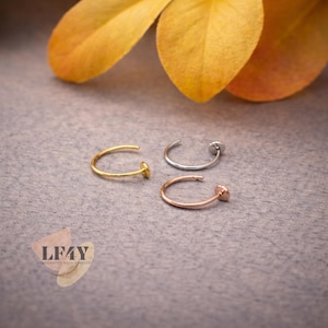 May include: Three nose rings in gold, silver, and rose gold, each with a small, round stud. The jewellery is displayed on a grey surface, with a blurred yellow leaf in the background. The rings are delicate and minimalist.
