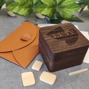 K&ouml;nnte beinhalten: Eine dunkle Holzbox mit der Gravur "LEAY HAND CRAFTED BODY JEWELLERY" liegt neben einem orangefarbenen Umschlag mit herzf&ouml;rmigem Verschluss. Mehrere kleine, rechteckige Holzst&uuml;cke sind daneben platziert.