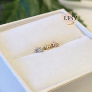 Puede incluir: Tres pequeños pendientes en forma de sol en plata, oro y oro rosa, cada uno con una gema transparente en el centro. Los pendientes se exhiben en una caja blanca, con la marca "LF4Y" visible en la esquina superior derecha.