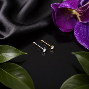 Könnte beinhalten: Zwei Nasenstecker, einer in Silber- und einer in Goldoptik, jeweils mit einem klaren, rund geschliffenen Edelstein in einer dreieckigen Fassung. Die Stecker haben einen geraden Stift mit einer kleinen Kugel am Ende auf dunkler Fläche.