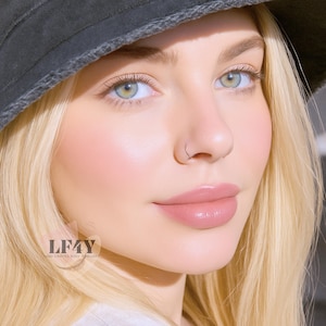 May include: A close-up of a person with blonde hair and a nose ring. The person has light skin, pink lips, and blue-green eyes. The image also shows a dark gray hat and the text "LF4Y HAND CRAFTED BODY JEWELLERY".