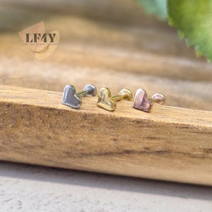 May include: Three heart-shaped stud earrings in silver, gold, and rose gold. The earrings are displayed on a wooden surface. The earrings are small and delicate, perfect for everyday wear. The LF4Y logo is visible in the upper left corner.