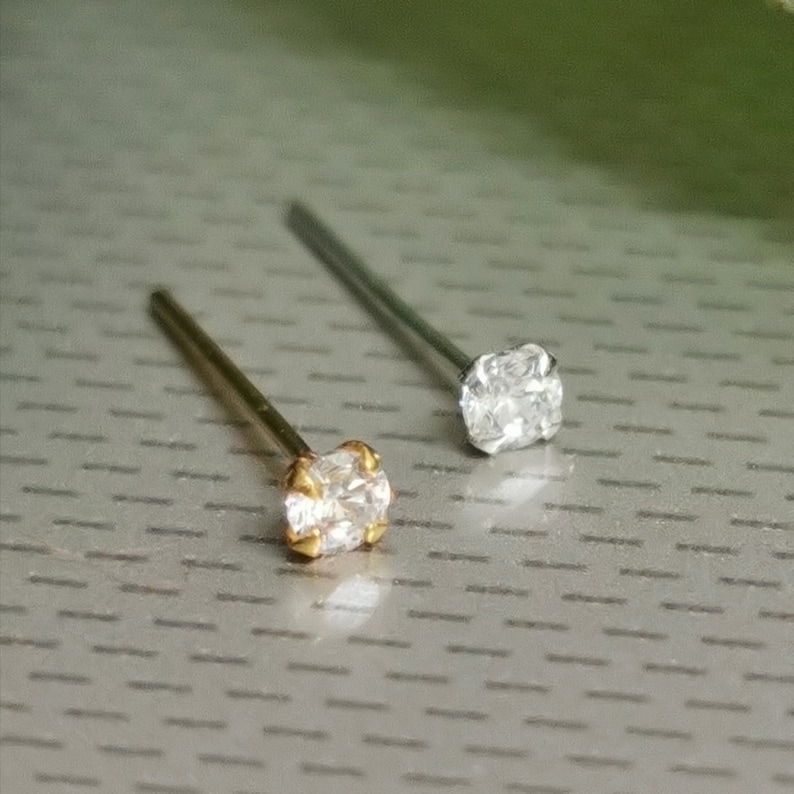K&ouml;nnte beinhalten: Zwei Nasenstecker. Einer hat einen goldfarbenen Stab und eine Fassung mit einem klaren, quadratisch geschliffenen Stein. Der andere hat einen silberfarbenen Stab und eine Fassung mit einem klaren, quadratisch geschliffenen Stein. Beide liegen auf einer grauen Oberfl&auml;che.
