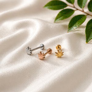 May include: Three ear studs with a cannabis leaf design. One stud is silver, one is rose gold, and the other is gold. The earrings are displayed on a white, textured surface with green leaves in the background.