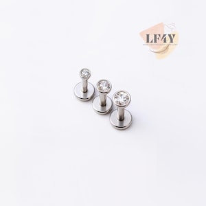 Può includere: Tre piercing per labbra color argento con base a disco piatto e un perno sormontato da una gemma trasparente, rotonda e scintillante. I piercing sono disposti in diagonale su sfondo bianco. L'immagine include il testo "LF4Y Hand Crafted Body Jewellery".