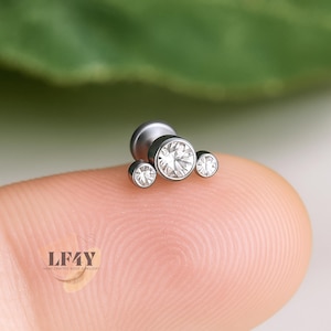 May include: A silver-toned body jewellery piece featuring three clear, round, sparkling gemstones. The largest gem is central, with two smaller gems either side. The jewellery is set against a blurred green background.