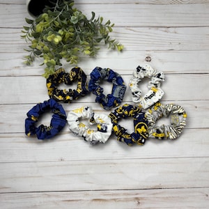 May include: A collection of seven fabric scrunchies in various patterns and colors, including navy blue, yellow, and white. Some scrunchies feature university-themed designs. The scrunchies are arranged on a light wood-grain surface.