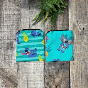 May include: Two small zippered pouches with a blue and green tropical print featuring Stitch from Lilo & Stitch. One pouch has a repeating pattern of Stitch with pineapples and stripes. The other pouch has a single image of Stitch wearing sunglasses and a lei.