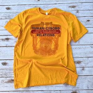 May include: A yellow t-shirt with a graphic of C-3PO from Star Wars. The text on the shirt reads "HUMAN-CYBORG FLUENT IN SIX MILLION FORMS OF COMMUNICATION RELATIONS".