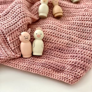 May include: Wooden farm animal figurines, including a pig, chicken, sheep, cow, and donkey, rest on a textured, dusky pink crocheted blanket. The figurines are simply painted and designed in a minimalist style. The blanket is a soft, muted pink.