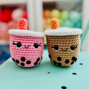 Könnte beinhalten: Zwei gehäkelte Boba-Tee-Plüschtiere, eines rosa und eines braun, mit schwarzen Boba-Perlen und lächelnden Gesichtern.