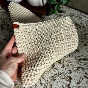 May include: Cream-colored, crocheted pot holder with a textured stitch pattern. A small, brown leather tab with a brass rivet is attached to the corner. The pot holder is folded in half, with a second one visible in the background.