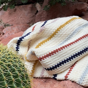 May include: A folded cream-coloured, hand-crocheted blanket with horizontal stripes in navy, mustard yellow, rust, and grey. The blanket displays a textured stitch and colour pattern. A cactus and red rocks are in the background.