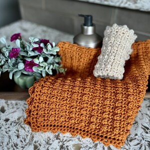 May include: Two crocheted dishcloths, one in a light beige and one in a burnt orange, are displayed on a countertop with a silver soap dispenser.