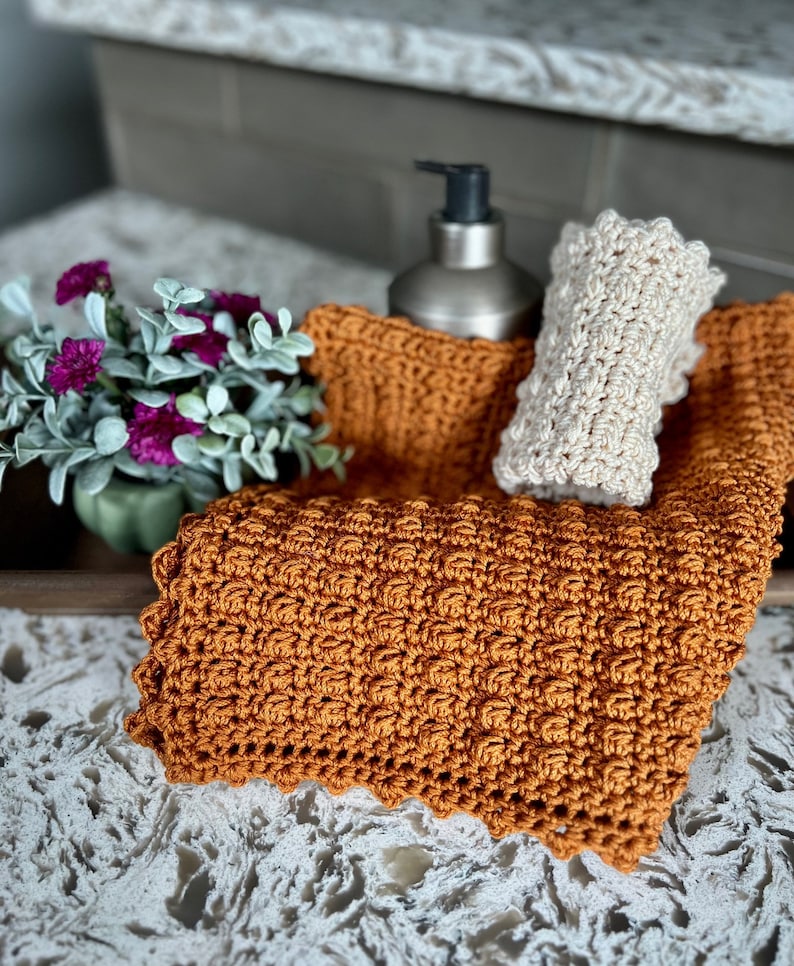 May include: Two crocheted dishcloths, one in a light beige and one in a burnt orange, are displayed on a countertop with a silver soap dispenser.
