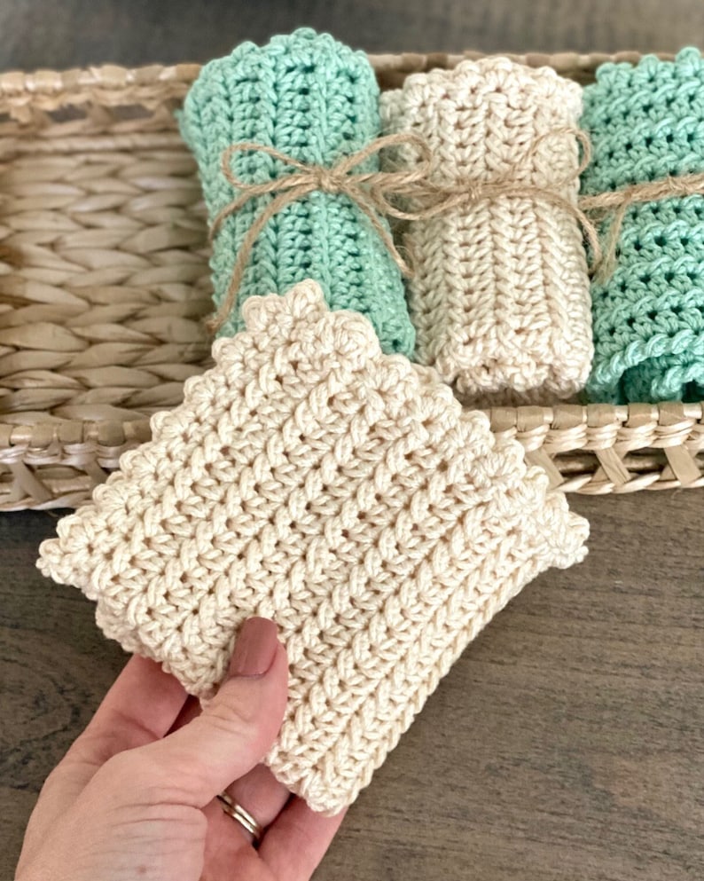 May include: A collection of handmade crochet washcloths in cream and seafoam green, tied with twine, and displayed in a woven basket. The washcloths have a textured pattern and scalloped edges. One cream washcloth is held in a hand.