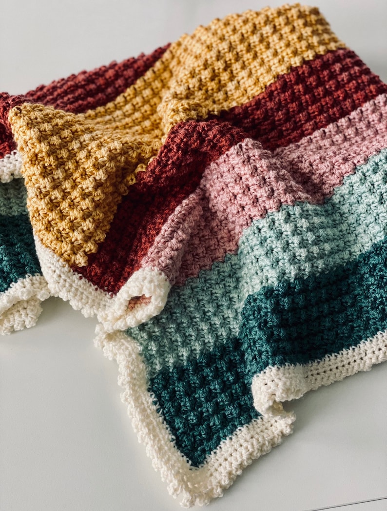 May include: A handmade, colorful crochet blanket with a textured stitch pattern. The blanket features horizontal stripes in shades of yellow, burgundy, pink, teal, and green, with a white scalloped border. The blanket is folded on a white surface.