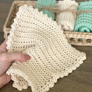 May include: A close-up of a beige crocheted washcloth with a scalloped edge. The washcloth is held in a hand, with other rolled washcloths in a wicker basket in the background. The washcloths are in shades of beige and mint green.