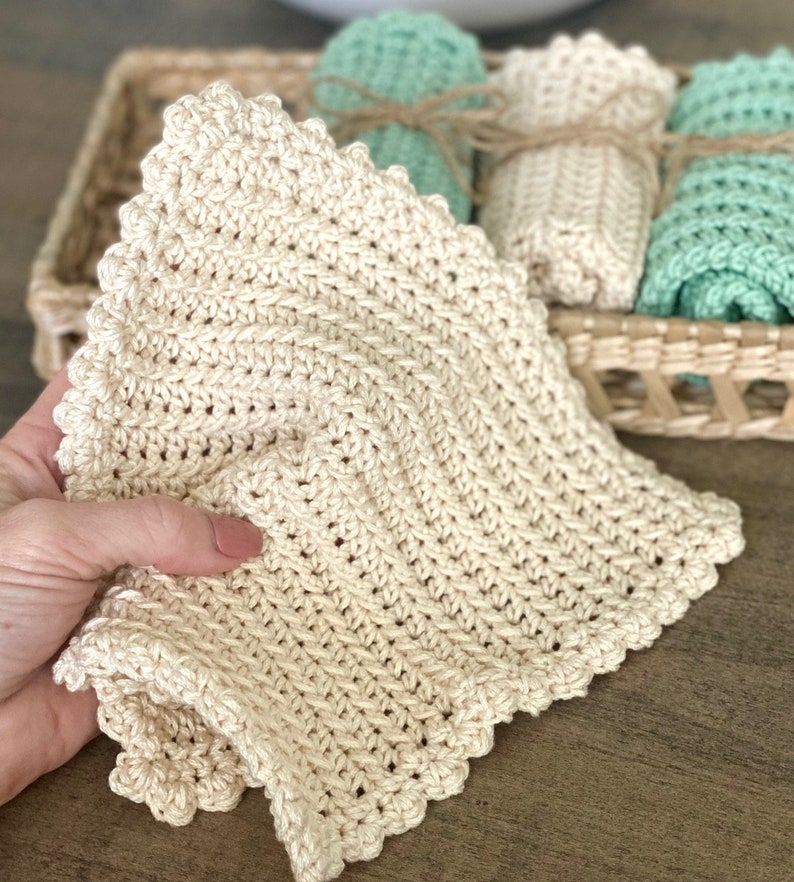 May include: A close-up of a beige crocheted washcloth with a scalloped edge. The washcloth is held in a hand, with other rolled washcloths in a wicker basket in the background. The washcloths are in shades of beige and mint green.