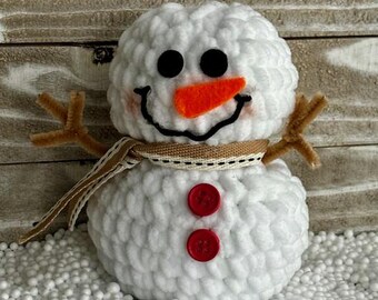 May include: A white crocheted snowman with a brown twig for arms, an orange felt nose, black felt eyes, and red buttons. The snowman is sitting on a bed of white styrofoam packing peanuts.