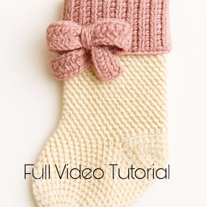 Pode incluir: Uma meia de Natal artesanal em creme e rosa empoeirado. A parte superior e o laço são de malha canelada em rosa empoeirado, enquanto o corpo é de malha creme. O texto "Full Video Tutorial" está na parte inferior.