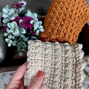 May include: Two crocheted dishcloths, one in a light beige color and one in a burnt orange color. Both dishcloths have a textured, raised pattern.