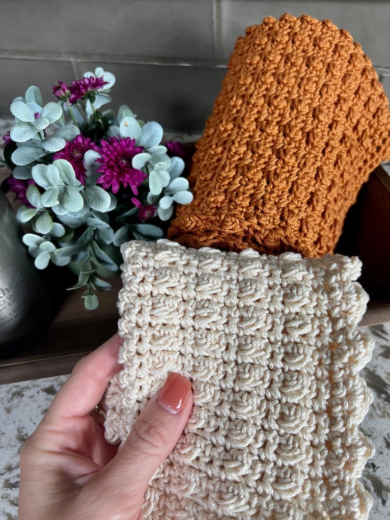 May include: Two crocheted dishcloths, one in a light beige color and one in a burnt orange color. Both dishcloths have a textured, raised pattern.