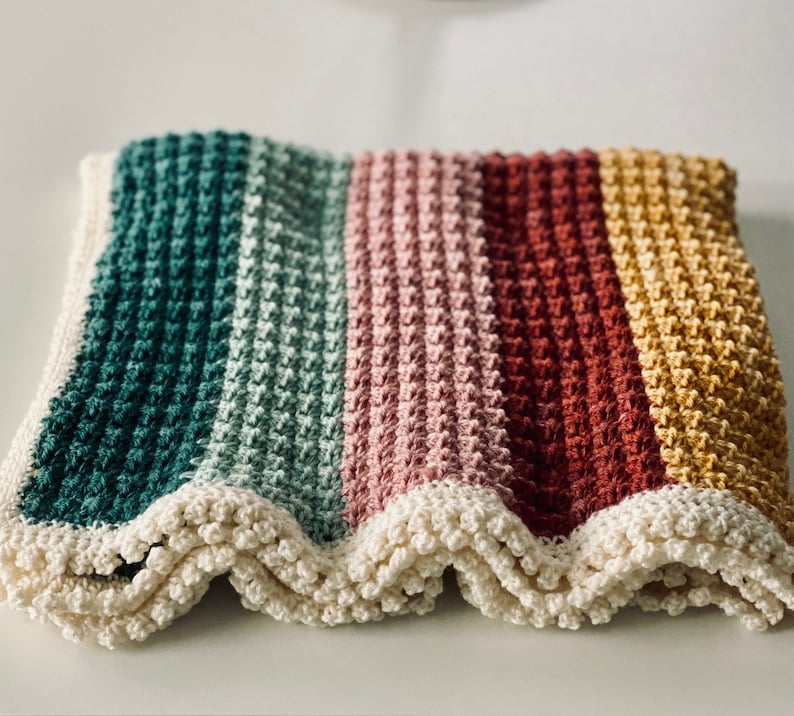 May include: A handmade crochet blanket with a striped pattern in teal, light green, pink, red, and yellow. The blanket has a scalloped, off-white border. The textured stitch creates a cozy, soft appearance.