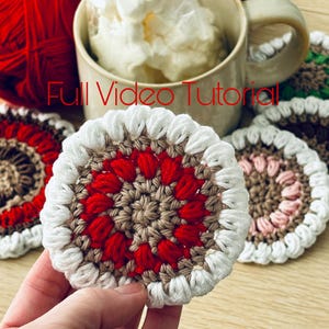 May include: A close-up of a handmade crochet coaster held in a hand. The coaster features a red, white, and tan floral design. A mug filled with a white substance and a ball of red yarn are in the background. The text "Full Video Tutorial" is visible.