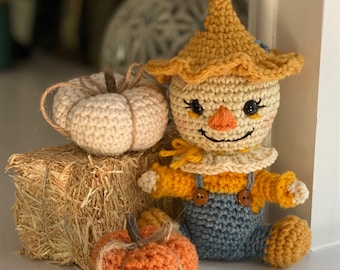 May include: A crocheted scarecrow doll with a yellow hat, blue overalls, and a friendly face. The doll is sitting next to two crocheted pumpkins, one white and one orange.
