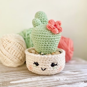Op de afbeelding: Een gehaakte cactus met een roze bloem, zittend in een witte gehaakte pot met een glimlachend gezicht. De cactus is groen en de pot heeft zwarte ogen en een zwarte mond.