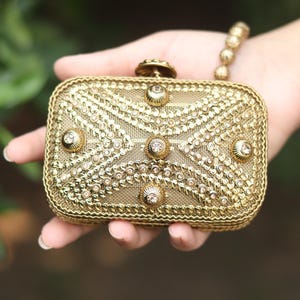 Op de afbeelding: Een rechthoekige gouden clutch met een ingewikkeld metalen gaasoppervlak versierd met rijen kleine heldere kristallen en vijf grotere ronde kristallen versieringen. De randen zijn afgewerkt met een gevlochten gouden bies en een sluiting.