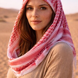 May include: A woman wearing a red and white checkered keffiyeh headscarf and a tan-colored garment. The headscarf is draped over the head and shoulders. The background is a desert landscape.