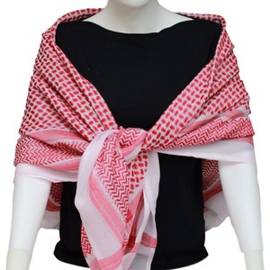 May include: A red and white checkered keffiyeh scarf draped over a mannequin. The scarf features a repeating pattern of red squares and diagonal lines. The scarf is wrapped around the neck and shoulders, with the ends tied in front.