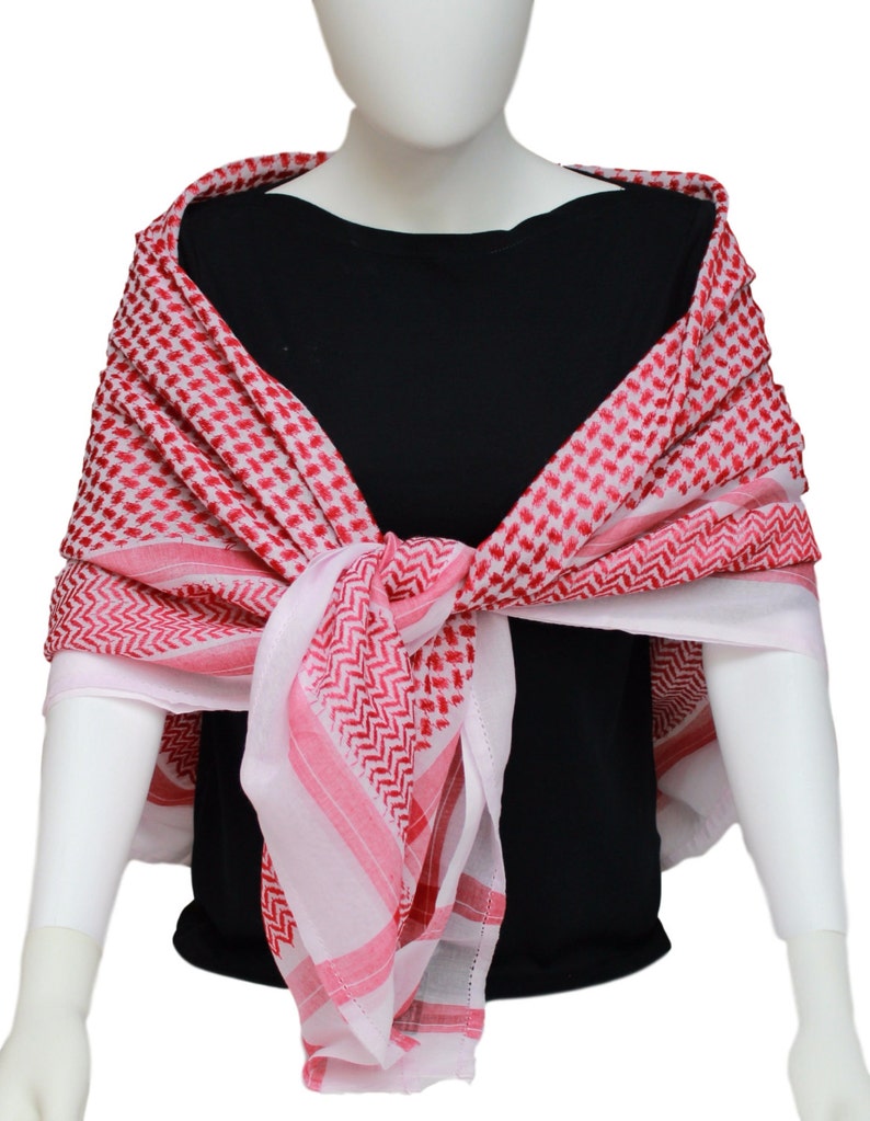 May include: A red and white checkered keffiyeh scarf draped over a mannequin. The scarf features a repeating pattern of red squares and diagonal lines. The scarf is wrapped around the neck and shoulders, with the ends tied in front.