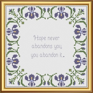 May include: Cross-stitch art in a gold frame. The design features a quote: "Hope never abandons you, you abandon it." The border includes purple flowers and green leaves on a white background.