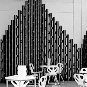 Könnte beinhalten: Schwarz-Weiß-Foto eines Café-Interieurs. Die Szene zeigt weiße Tische und Stühle mit einem einzigartigen Design vor dem Hintergrund einer dekorativen Wand mit einem sich wiederholenden Muster aus vertikalen Elementen.