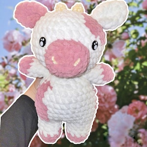 May include: A crocheted plush toy cow with pink and white fur. The cow has a pink nose and black eyes. The toy is sitting on a hand.