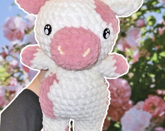 May include: A crocheted plush toy cow with pink and white fur. The cow has a pink nose and black eyes. The toy is sitting on a hand.