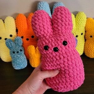 May include: A collection of colourful crocheted bunny plushes in various colours, including pink, yellow, blue, orange, and light blue. The plushes are all shaped like bunnies with long ears and are made with a soft, textured yarn.